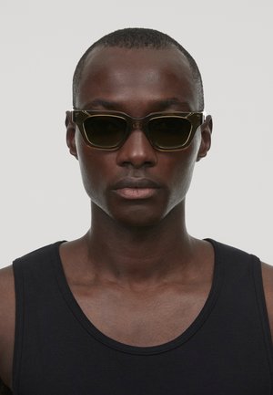 Schwarz gerahmte Sonnenbrille mit dunklen Gläsern, die eine einzigartige geometrische Form und einen kantigen Design aufweisen. Getragen mit einem schwarzen Trägershirt.