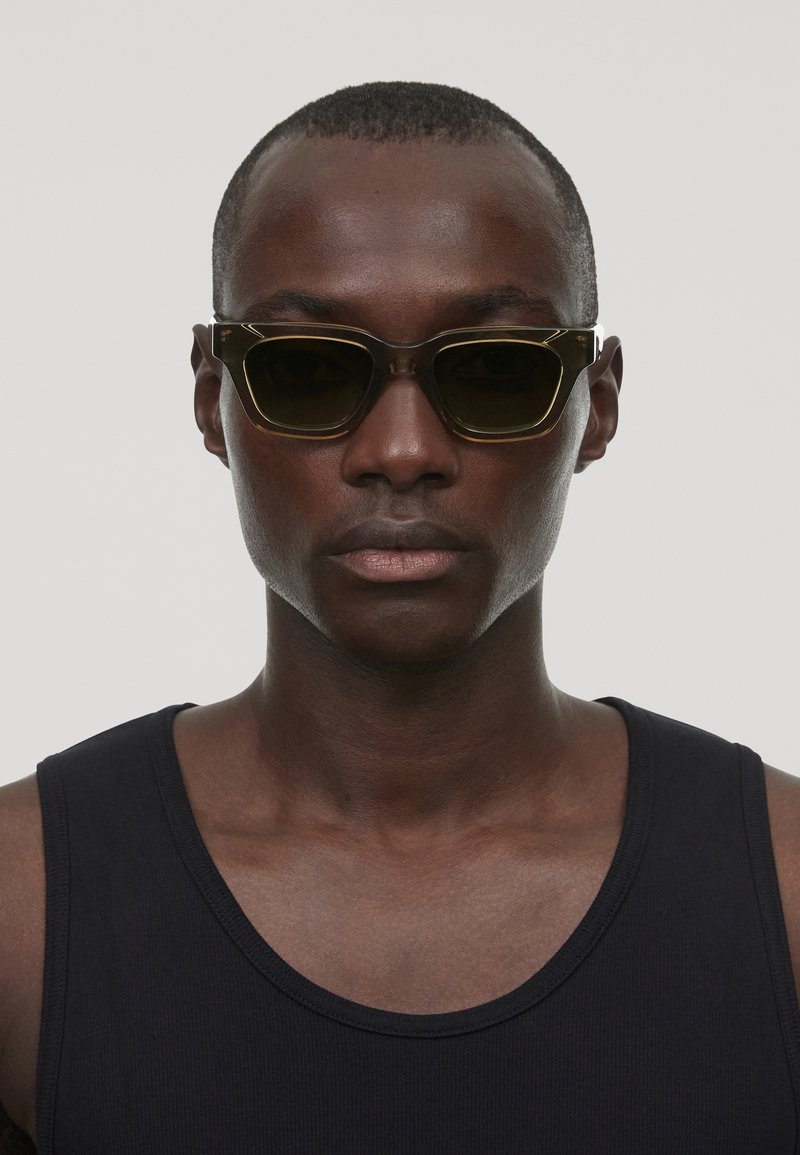 Lunettes de soleil à monture noire avec verres sombres, présentant une forme géométrique unique et un design angulaire. Portées avec un débardeur noir.