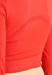 Červený sportovní cropped top s hladkou texturou, zaobleným výstřihem a elastickým pruhem s logem "STELLA/MCCARTNEY" na lemu.