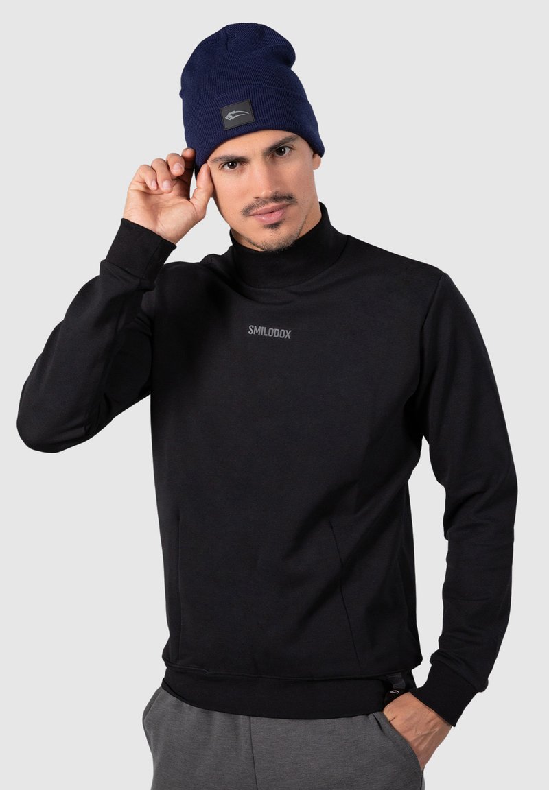 Felpa nera con collo alto, texture morbida e logo con marchio. Abbinata a un berretto lavorato a maglia blu, con un piccolo logo a toppa.