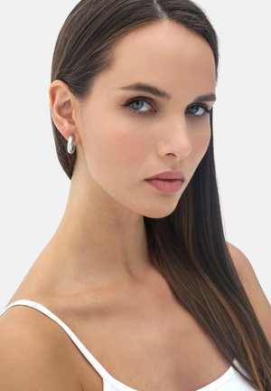 Giovane donna con capelli lunghi e scuri che indossa una canottiera bianca e orecchini a cerchio argentati, che guarda leggermente di lato su uno sfondo semplice.