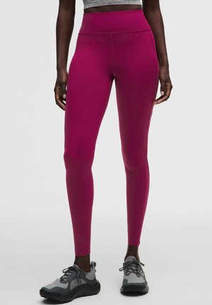 Kvinde iført lyserøde høj-talje leggings og grå træningssko, stående med arme afslappet ned langs siden på en ensfarvet hvid baggrund.