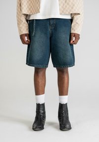 Shorts en denim bleu foncé avec une coupe décontractée, présentant des ourlets cousus. Portés avec des chaussettes blanches et des bottines noires.