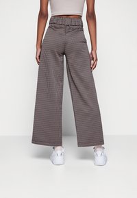 Hnědé a bílé kalhoty s širokými nohavicemi ve vzoru houndstooth s elastickým pasem, spárované s bílými teniskami na jednobarevném pozadí.