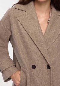Kobieta ubrana w taupe'owy, podwójnie zapinany płaszcz wełniany z szerokimi klapami i zapiętymi mankietami, z ręką w kieszeni, na białym tle.