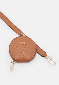 Okrągła brązowa portmonetka na monety z ekoskóry o teksturowanej powierzchni, złotymi akcentami i odpinanym paskiem. Z przodu znajduje się logo "GUESS".