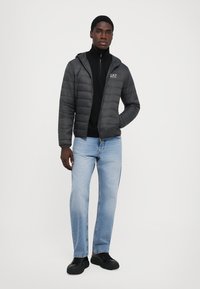 Veste noire matelassée avec capuche, logo gris, portée sur un col roulé noir, associée à un jean bleu clair et des baskets noires.