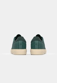 Green suede sneakers with a low-top design and cream rubber soles. Features a textured finish and minimalist style with elastic accents.