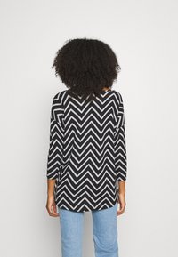Top con motivo a zigzag bianco e nero con maniche a tre quarti, scollo rotondo e orlo posteriore leggermente più lungo, abbinato a jeans azzurri chiari.