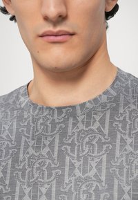 Szary t-shirt z grafiką, z powtarzającym się wzorem logo, okrągłym dekoltem i gładką fakturą. Tkanina wydaje się lekka i casualowa.