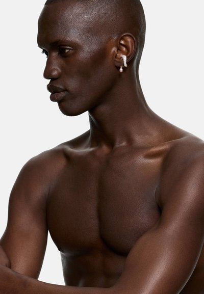 Side profile of a shirtless man with dark skin wearing a silver and pearl earring on his left ear against a plain background.