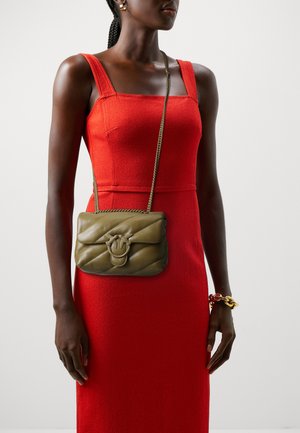 Borsa a tracolla in pelle verde oliva con motivo trapuntato, dotata di chiusura a corda intrecciata. Il modello indossa un vestito rosso senza maniche, mostrando la borsa.