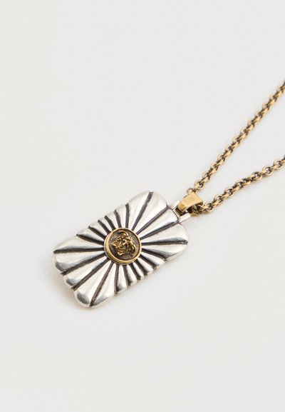 Pendentif rectangulaire en argent avec des lignes rayonnantes et un emblème de tête de lion doré sur une chaîne en or.