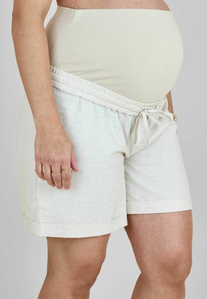 Personne enceinte portant un short de maternité beige avec une taille ajustable par-dessus un bandeau de soutien ventre couleur chair, debout sur un fond uni.