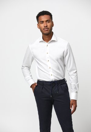 Witte blouse met lange mouwen en een knopenlijst aan de voorkant met gouden knopen, gecombineerd met marineblauwe broek met trekkoord, die een getailleerde pasvorm laat zien.