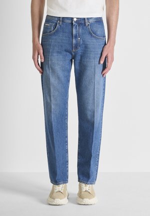 Persoon draagt rechtgesneden blauwe spijkerbroek en beige sneakers, staand tegen een effen witte achtergrond.