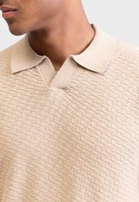 Polo en tricot beige avec un motif en losanges texturé, doté d'un col en V et d'un col doux. Le tissu semble léger et respirant.