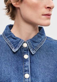 Τζιν πουκάμισο με δομημένο γιακά, ασημένια κουμπιά τύπου snap και αντίθετες ραφές. Το ύφασμα έχει μεσαίο μπλε χρώμα με ομαλή υφή.