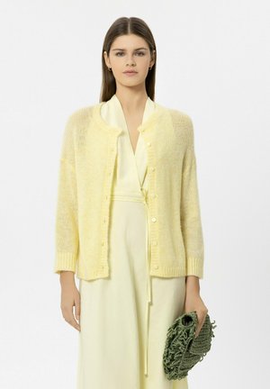 Junge Frau in hellgelbem Kleid und passendem weichem Strickcardigan, die eine strukturierte grüne Clutch vor weißem Hintergrund hält.