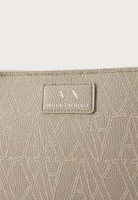 Bež torba od teksturirane kože sa geometrijskim uzorkom i ugraviranom oznakom s logotipom "A/X" i "ARMANI EXCHANGE" u srebrnoj boji.