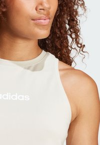 Top atlético sin mangas de color gris claro con un escote redondo, que presenta un prominente logo blanco de Adidas en la parte frontal. Material suave y elástico.