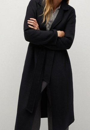 Mujer con cabello largo que lleva un abrigo negro con cinturón sobre un suéter gris, de pie con los brazos cruzados frente a un fondo liso.