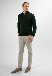 Man in een donkergroene kabeltrui, beige broek en brown instapschoenen, staand met handen in zijn zakken tegen een effen achtergrond.