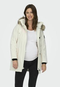 Parka bianca con cappuccio foderato in pelliccia, chiusura con zip e bottoni, tasche frontali e dettagli neri sui polsini, indossata sopra una maglietta bianca e jeans neri.