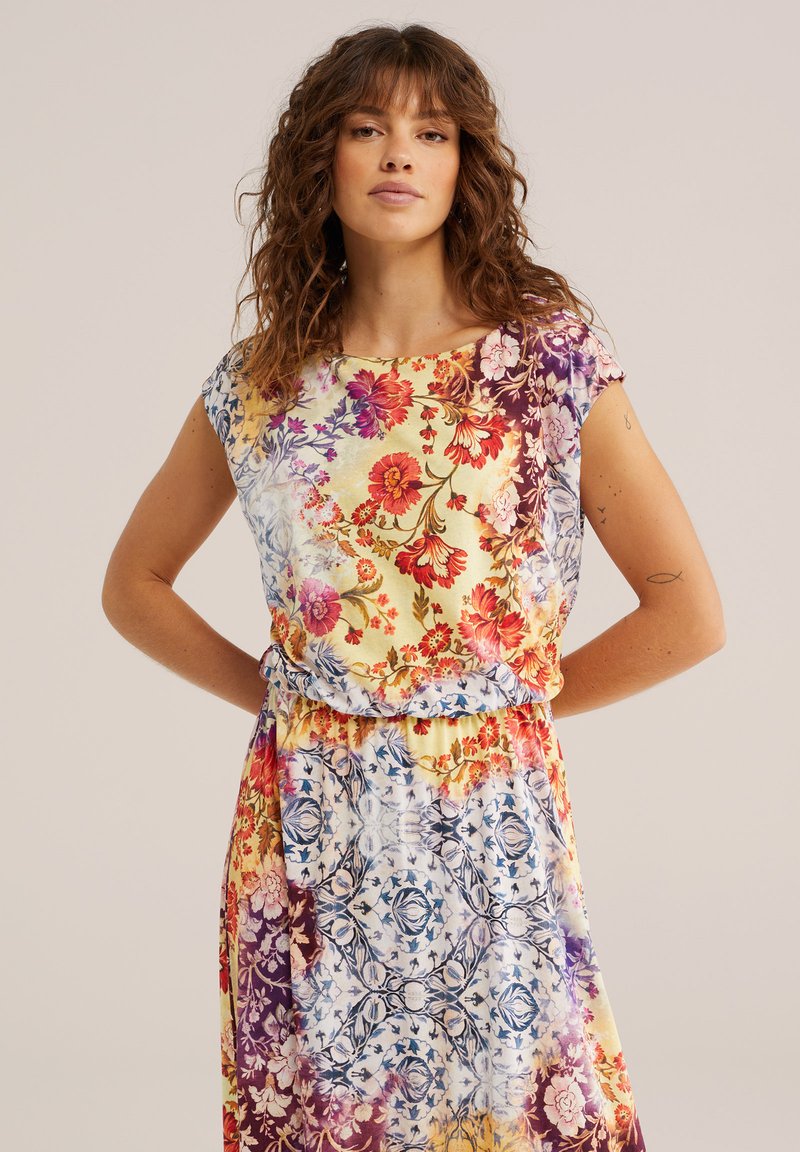 Frau mit lockigem braunem Haar, die ein farbenfrohes, blumiges Kleid mit roten, gelben, lila und blauen Mustern trägt und mit den Händen in den Hüften vor einem einfarbigen Hintergrund steht.