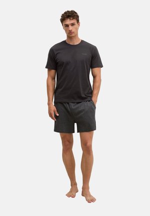 Junger Mann steht barfuß, trägt ein schwarzes T-Shirt mit kurzen Ärmeln und schwarze karierte Shorts vor einem weißen, einfarbigen Hintergrund.