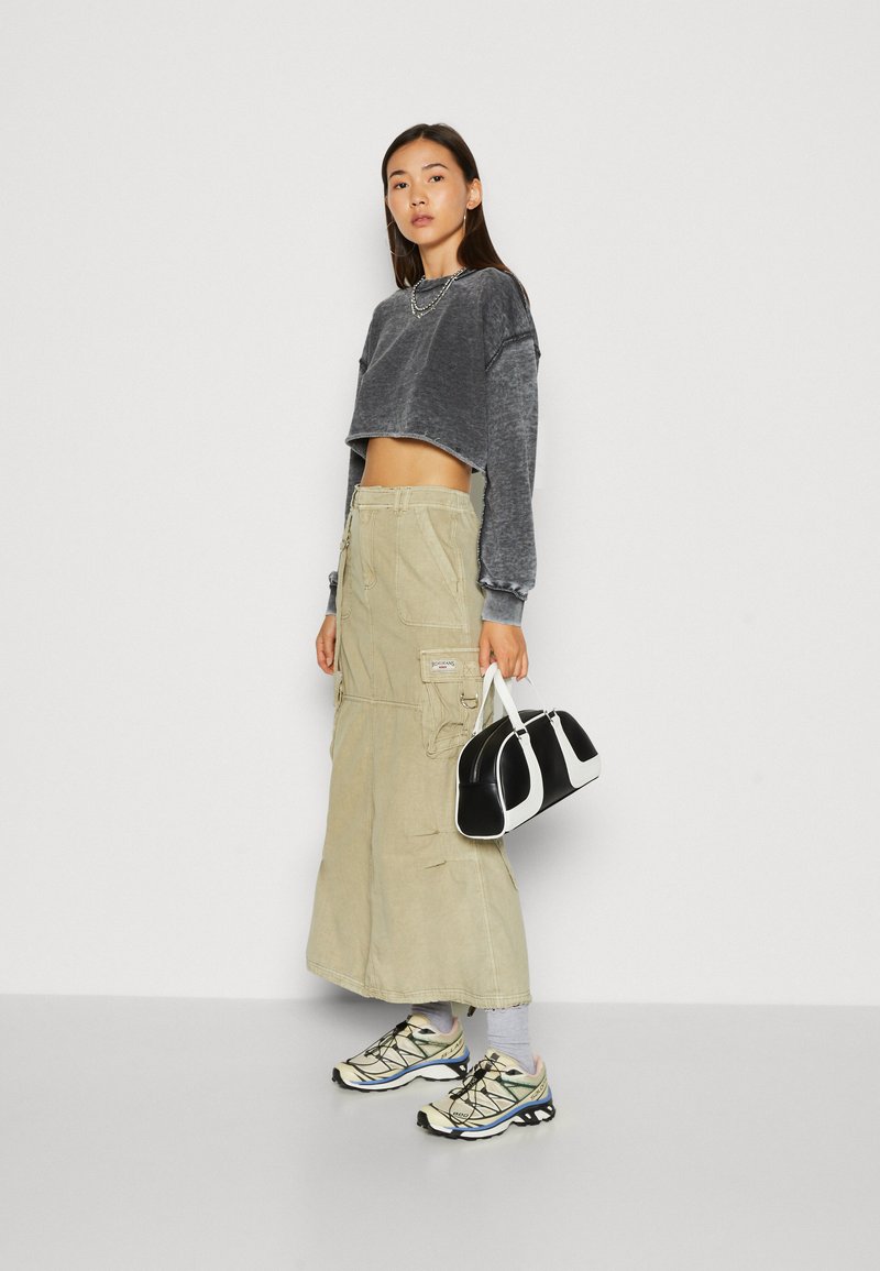 Felpa grigia corta, gonna cargo beige e sneakers sportivi beige e neri. La modella porta una piccola borsa a tracolla bianca e nera.