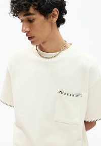 Krátky rukáv, off-white mikina z mäkkej látky, obsahujúca náprsné vrecko s čiernym prešívaním a hladký výstrih.