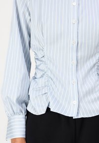 Lyseblå stripete bluse med samlet sidesøm, knapp-down front og lange ermer med knappelister. Stoffet har en glatt tekstur.