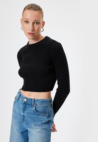 Zwarte cropped gebreide trui met geribbelde textuur en kabelpatroon, gecombineerd met high-waisted lichtblauwe spijkerbroek met knoopsluiting en zakken.
