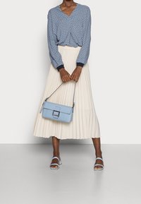 Frau in einer gemusterten blauen Bluse und einem cremefarbenen, plissierten Midikleid, die eine hellblaue Handtasche und hellblaue Absatzsandalen hält und vor einer weißen Wand steht.