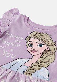 Lila bomulls t-shirt med en grafik av en karaktär med flätat hår. Volangärmar och texten "Omfamna magin i dig" visas.