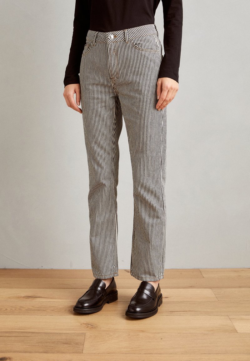 Stripete svarte og hvite bukser med en tett livvidde, fem lommer og et rett snitt, kombinert med brune loafers på et tregulv.