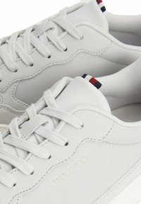 Białe skórzane sneakersy o gładkiej fakturze, z tonalnym przeszyciem, sznurowanym zapięciem oraz trójkolorową naszywką z tyłu. Wytłoczone logo „HILFIGER”. 