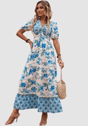 Vestido maxi floral en blanco con patrón de flores azules, mangas cortas abullonadas, detalle de encaje y diseño en capas. Accesorizado con un bolso de mimbre.