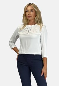 Biała, długosleeve bluzka z plisowanym dekoltem, w połączeniu z granatowymi spodniami w kropki. Materiał wydaje się gładki i lekki.