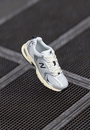 Gray and white running shoe with mesh upper, rubber sole, and thick cushioning, featuring 'N' logo and laces. Positioned on a textured surface.