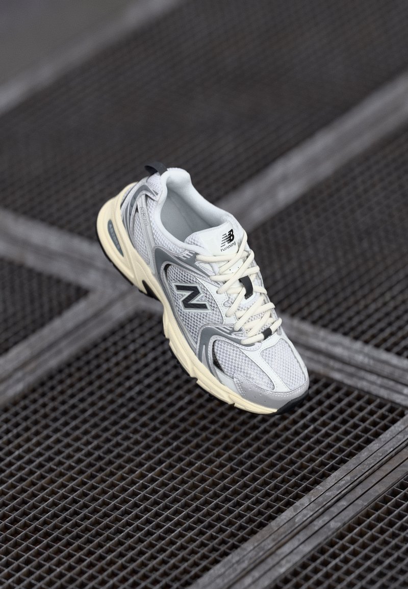 Scarpa da corsa grigia e bianca con tomaia in mesh, suola in gomma e spessa imbottitura, caratterizzata dal logo 'N' e dai lacci. Posizionata su una superficie texture.