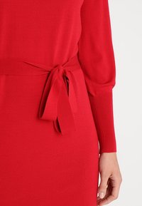 Robe tricotée rouge avec une silhouette ajustée, un lien à la taille et des détails de manches côtelées. Texture lisse et couleur vive.
