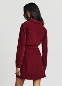 Vestito in velluto a coste rosso con maniche lunghe, colletto e dettaglio con cintura. Presenta una texture a coste verticali e una silhouette aderente con orlo svasato.