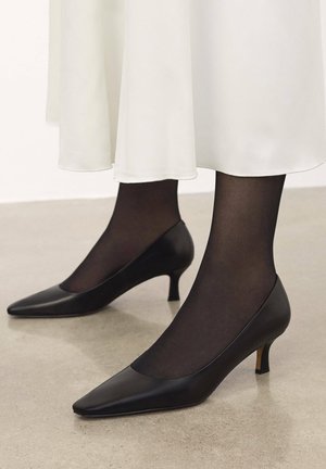 Scarpe nere in pelle con tacco alto e punta affilata, con un tacco kitten basso, indossate con calze nere trasparenti, sotto un abito bianco fluente.
