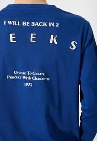 Blaue Langarmbluse mit weißem Text "ICH WERDE IN 2 WOCHE ZURÜCK SEIN" und kleinerem Text "ENTSCHEIDE DICH, EIN PRODUKT MIT CHARAKTER ZU CREIEREN 1972"