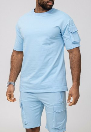Homme portant une chemise à manches courtes bleu clair et un short cargo assorti avec des poches à rabat sur la manche et le short, montre-bracelet au poignet gauche.