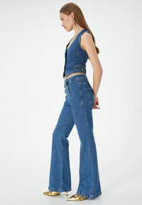 Denimväst och utsvängda jeans i mellanblått, med en figurnära design, knappdetalj och slät textur. Gyldene spetsiga skor kompletterar stilen.