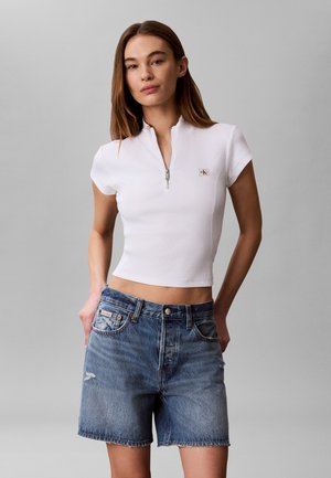 Kvinde iført en hvid kortærmet crop-top med lynlås og blå cowboyshorts med hænderne i lommerne mod en ensfarvet grå baggrund.