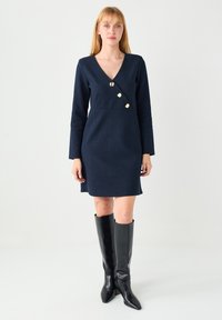 Robe portefeuille bleu marine à manches longues, en tissu texturé, ornée de boutons dorés et d'un décolleté en V. Portée avec des bottes en cuir noir montant jusqu'au genou.